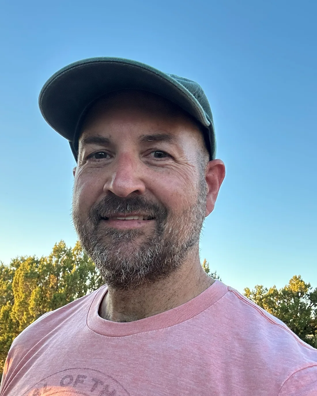 Photo of Jonathan Sandmel smiling outdoors at golden hour, wearing a green cap, with New Mexico juniper hills behind him.