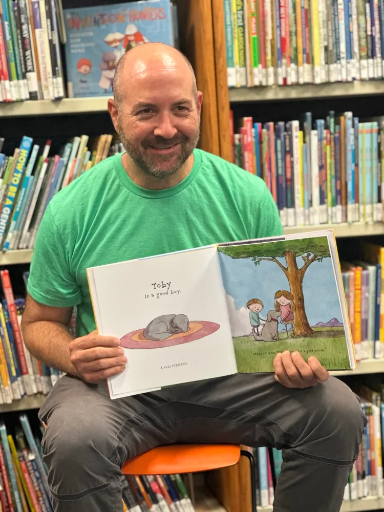 Jonathan Sandmel, smiling in a green t-shirt in a library, holds open Toby's a Mensch to a spread of Toby curled up on a rug.